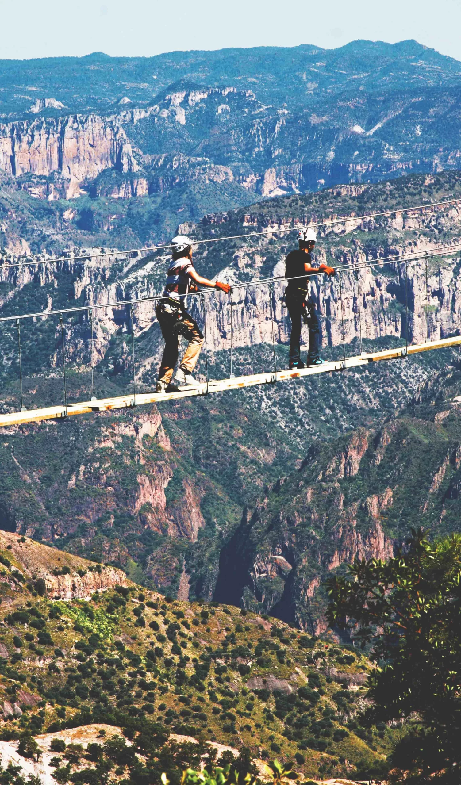 Ecoproparks. Dos niños con equipo de protección disfrutando de una emocionante aventura sobre un puente de madera a gran altura, con impresionantes vistas de las majestuosas montañas en Ecoproparks.